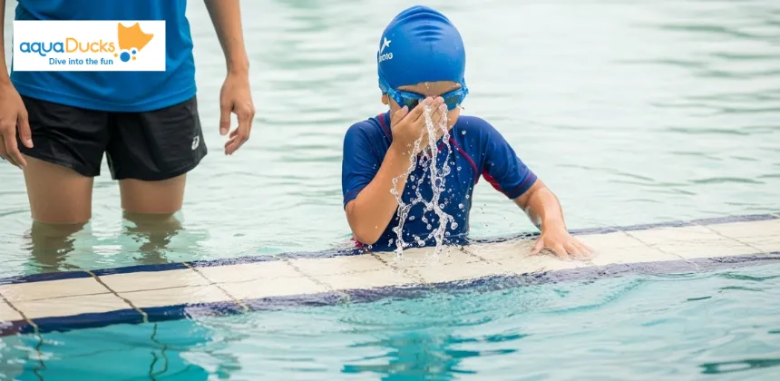 A young beginner swimmer wearing goggles and a swim cap gently splashes water on his face near the pool edge to build water confidence before learning how to swim.