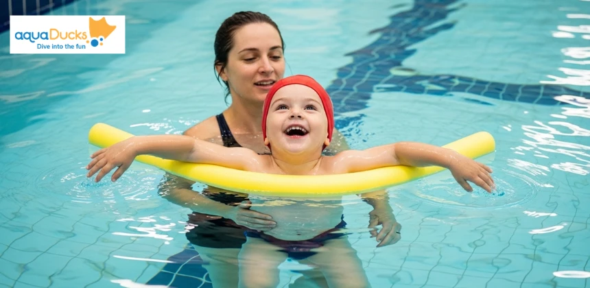 A joyful toddler floating confidently with a pool noodle and instructor support during a playful swimming lesson in a bright indoor pool 
