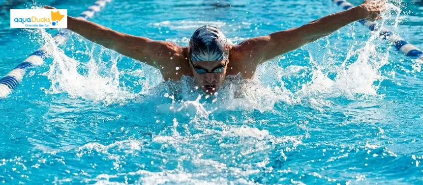 A swimmer performing butterfly stroke in an outdoor pool, both arms extended outward during recovery phase, water splashing on both sides