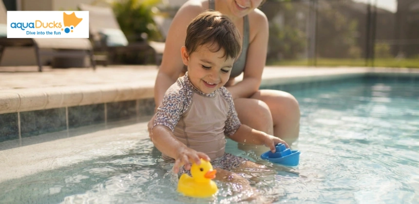 A young boy smiling and playing with a rubber duck and a blue toy boat in a swimming pool while sitting beside his mother.