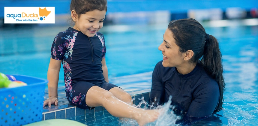 A young girl smiling as she splashes her feet in the pool while an instructor looks on encouragingly.