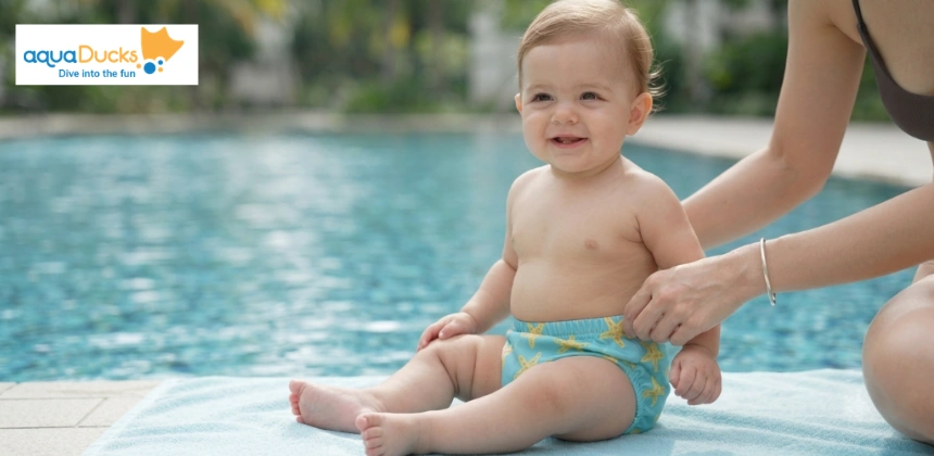 A happy baby in a teal starfish-print swim diaper sits on a towel by a pool, with a parent's hands nearby