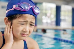 Can You Swim With an Ear Infection? Young swimmer holding her ear in pain by the pool edge during swim lessons