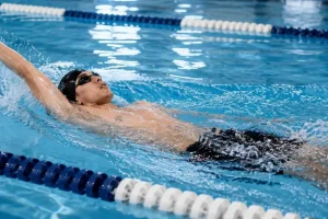 A man performing the backstroke swimming style in a lap pool with lane ropes, showing proper arm and head position