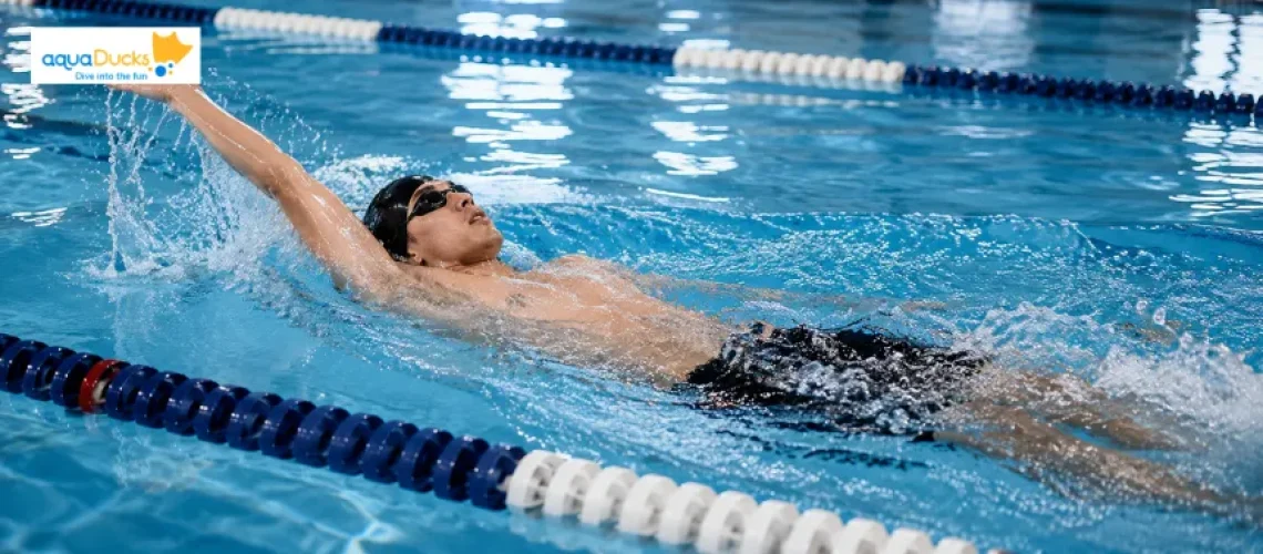 A man performing the backstroke swimming style in a lap pool with lane ropes, showing proper arm and head position