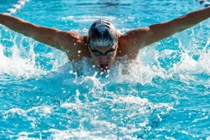 A swimmer performing butterfly stroke in an outdoor pool, both arms extended outward during recovery phase, water splashing on both sides
