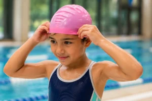 A young child adjusting a pink silicone cap while learning how to wear a swimming cap properly at an indoor pool.